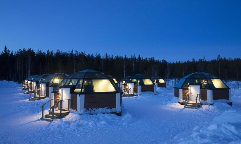 Row of glass igloos at Arctic SnowHotel & Glass Igloos at dusk, with lit glass roofs and snow on the ground