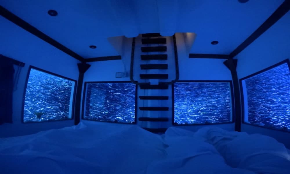 Bedroom of the original Manta Resort Underwater Room at night, with lit windows on four sides; this room was replaced in May 2026